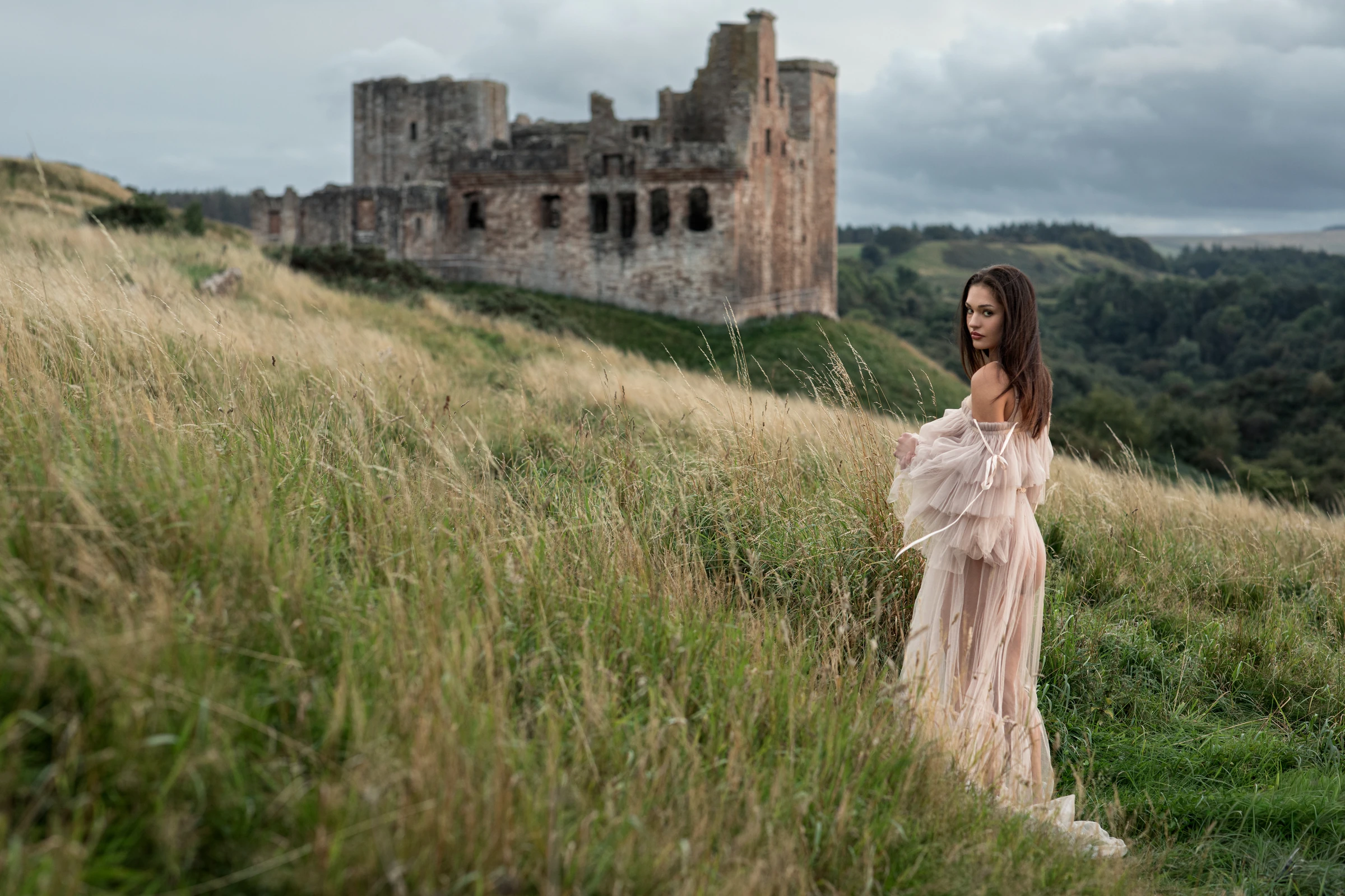 Editorial portrait of a woman in a tulle gown in long grass below castle ruins in Scotland