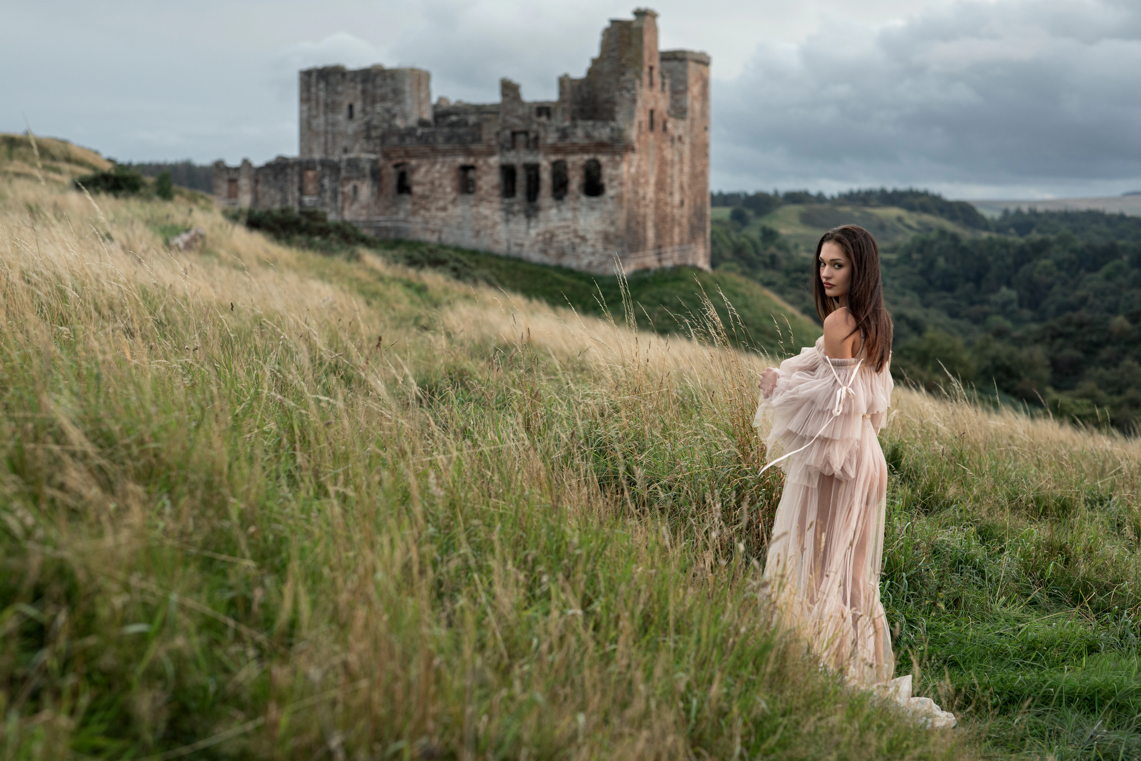 Editorial portrait of a woman in a tulle gown in long grass below castle ruins in Scotland