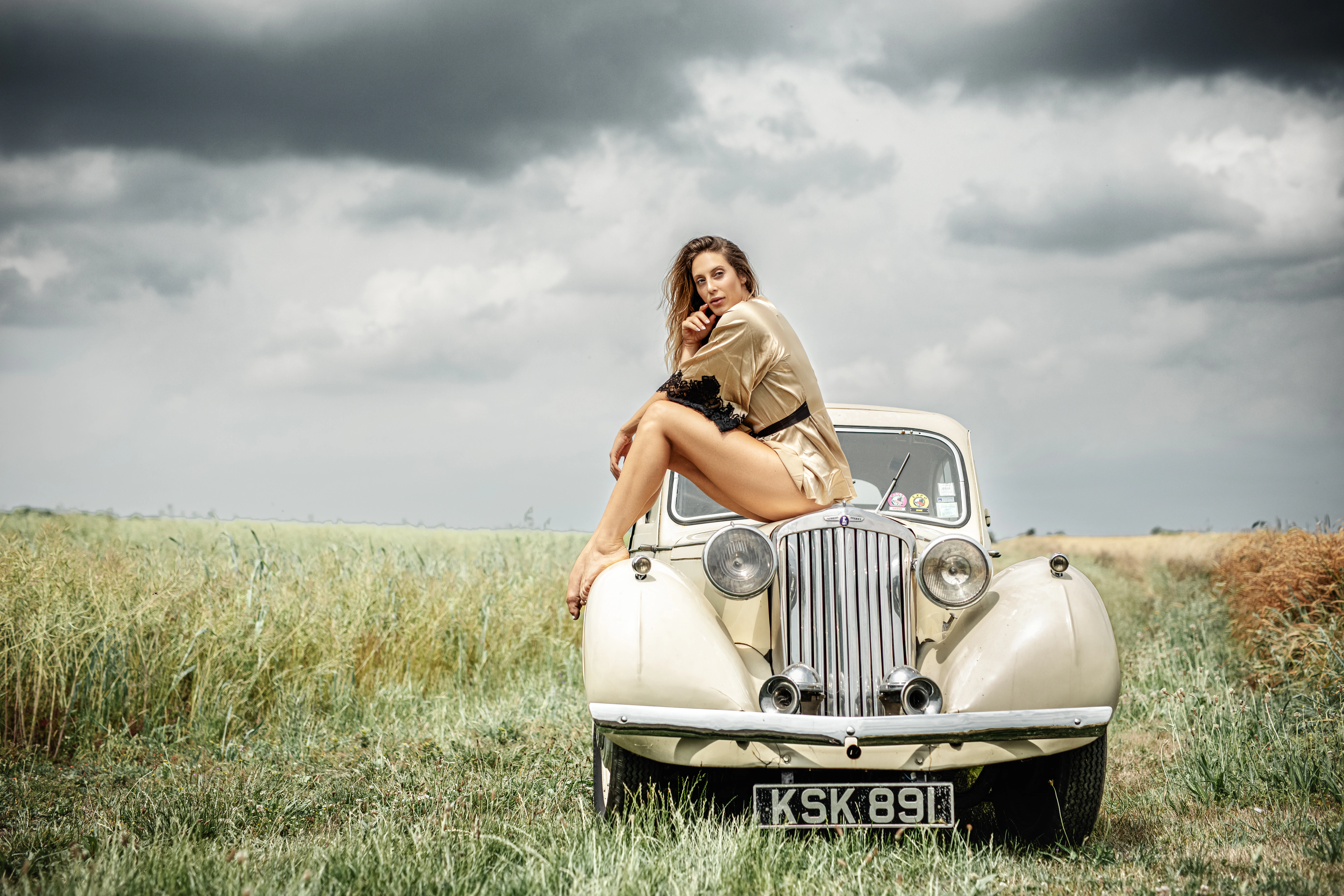 Editorial portrait of a woman sitting on a vintage car in a wild field under a stormy sky